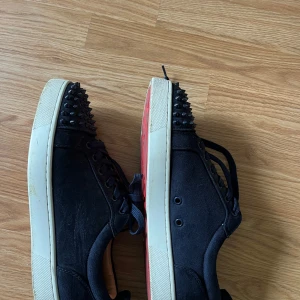 Mörkblå loubs - Säljer ett par svarta sneakers från Christian Louboutin med svarta nitar på tån och klassisk röd sula. Skorna har snörning och en platt vit sula. Perfekta för dig som vill sticka ut med en edgy stil.