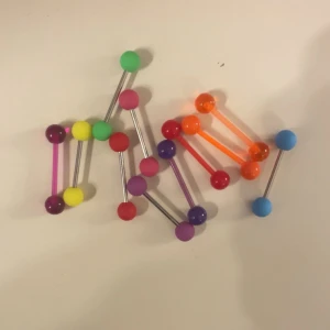 Färgglada piercingstavar med kulor - Säljer flera piercingstavar i olika färger med kulor i plast och metall. Färgerna inkluderar lila, gul, grön, röd, rosa, blå och orange. Perfekt för dig som vill variera din stil med färgglada detaljer.