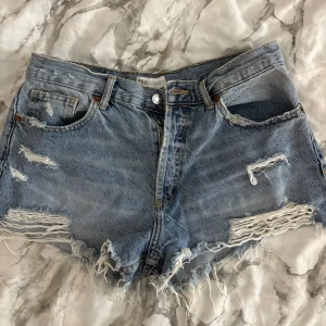 Blå jeansshorts med fransar från Zara - Säljer ett par blå jeansshorts från Zara med råa kanter och slitningar framtill. Sitter jätte skönt på. Perfekta för sommaren!