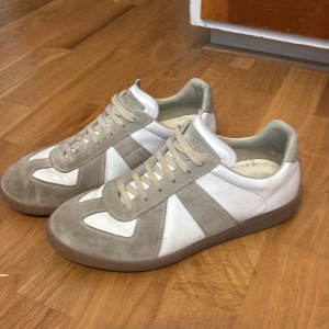 Beige och vita sneakers med mockadetaljer - Snygga sneakers i beige och vitt med paneler i mocka och skinn. Skorna har snörning och en platt sula, perfekt för en stilren vardagslook. Klassisk design som passar till det mesta.