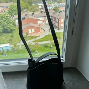 Svart axelväska - Säljer en stilren svart axelväska med minimalistisk design. Väskan har både ett långt och ett kort handtag, vilket gör den enkel att bära på olika sätt. Perfekt för dig som gillar klassiska accessoarer med modern touch. Axelremmen är avtagbart. Knappt använd. 