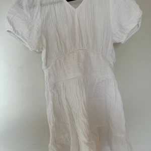 Vit kortärmad klänning från Gina Tricot - Säljer en söt vit klänning från Gina Tricot i storlek 146/152. Klänningen har korta puffärmar, v-ringning och smockdetaljer i midjan. Tillverkad i ett lätt och luftigt material, perfekt för varma dagar.