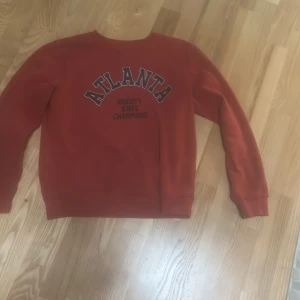 Röd sweatshirt med Atlanta-tryck från 157 - Säljer en röd sweatshirt från 157 med trycket 'ATLANTA VARSITY STATE CHAMPION' på bröstet. Tröjan har rund hals och långa ärmar, perfekt för en avslappnad stil. Använd få tal gånger