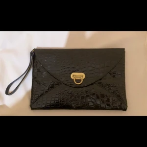 Svart kuvertväska med krokodilpräglat mönster - Stilren svart kuvertväska i syntet med krokodilpräglad yta och guldfärgat spänne framtill. Enkel och elegant design.