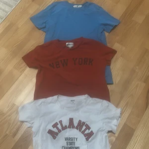 Tre t-shirts i olika färger och tryck - Säljer tre t-shirts: en vit med 'ATLANTA VARSITY STATE CHAMPIONS'-tryck, en röd med 'NEW YORK'-tryck och en enkel blå t-shirt. Alla har rund hals och korta ärmar. Perfekta för en avslappnad stil. Det är olika ställen tröjorna är ifrån. 60kr för alla tillsammans 