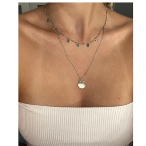 Dubbel silverhalsband med berlocker och hjärta - Snyggt dubbelt halsband i silverfärg. Det ena lagret har små droppformade berlocker och det andra ett runt hjärthänge. Perfekt för att ge en trendig touch till din outfit.