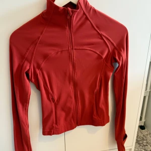 Röd träningsjacka från Lululemon - Säljer en röd, figurnära träningsjacka från Lululemon. Jackan har långa ärmar och är tillverkad i ett stretchigt syntetmaterial som passar perfekt till träning. köpt ny och bara använd 1-2 gånger. säljer då den inte kommer till användning 