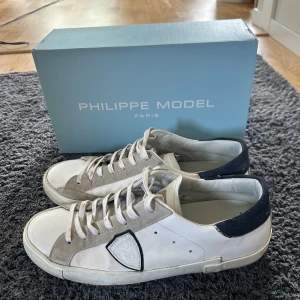 Philippe model - Snygga vita sneakers från Philippe Model med grå mockadetaljer och mörkblå häl.  Perfekta för en stilren vardagslook. Skorna levereras med originalkartong och dustbag. Digitalt kvitto kan även visas innan köp. Skorna är i fint skick utan några synliga defekter. Nypris ca 4000. Mitt pris 2200, kan diskuteras.