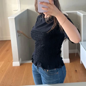 Svart smockad topp - Säljer en svart topp med smockad effekt över hela kroppen. Toppen har korta ärmar och en figurnära passform som framhäver midjan. Perfekt att styla med jeans eller kjol.
