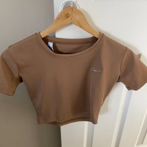 Beige croppad tränings tröja från Nike - Säljer en beige croppad t-shirt från Nike med klassisk rund halsringning och korta ärmar. Diskret Nike-logga broderad på bröstet. Perfekt för en sportig och avslappnad stil.