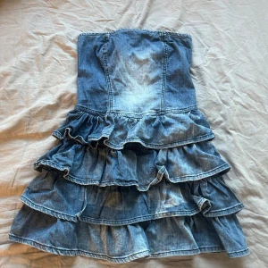 Jeansklänning med volanger från Gina Tricot - Säljer en snygg axelbandslös jeansklänning från Gina Tricot i storlek XS. Klänningen har flera lager volanger och dragkedja bak. Perfekt för dig som gillar denim och vill sticka ut med en unik look.