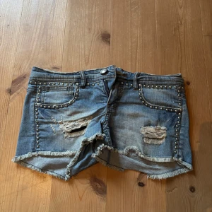Jeansshorts med nitar och fransar - Snygga blå jeansshorts med slitningar, fransig kant och silverfärgade nitar längs fickor och linning. Klassisk femficksmodell med låg midja och råa detaljer för en cool look.