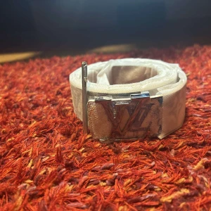 Vit Bälte med monogram - Säljer ett stilrent vitt bälte med klassiskt monogram-mönster och ett silverfärgat spänne. Bältet är smalt och har flera hål för justering. Perfekt accessoar för att lyfta din outfit.