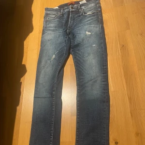 Jack&Jones Slimfit/Glenn  - Säljer ett par blå slim fit jeans från Jack & Jones med slitna detaljer och klassisk femficksdesign. Jeansen har normal midja och lätt tvättad look, perfekta till vardags. Modellen heter Glenn. Helt oanvända, skick: 10/10, storlek: Waist - 29, Length - 30