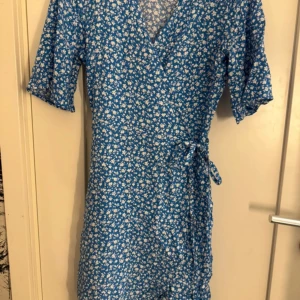 Blå blommig omlottklänning - Säljer en blå omlottklänning med vitt blommönster och knyt i midjan. Klänningen har v-ringning och korta ärmar, perfekt för varma dagar. Lätt och luftig modell som ger en somrig känsla.