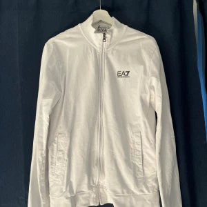 Vit zip-jacka från EA7 Emporio Armani - Säljer en stilren vit jacka från EA7 Emporio Armani med dragkedja framtill och diskret logga på bröstet. Jackan har långa ärmar, ståkrage och två fickor med dragkedja. Perfekt för en clean och sportig look.
