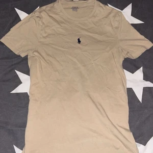 Beige t-shirt från Polo Ralph Lauren - Säljer en beige t-shirt från Polo Ralph Lauren med klassisk broderad logga på bröstet. T-shirten har rund halsringning och är i en slim fit-modell. Perfekt för en stilren och enkel look.