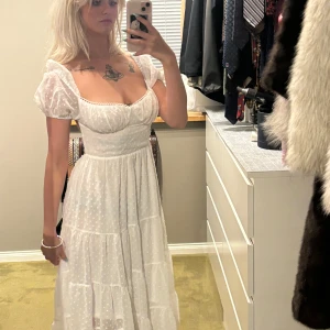 Romantisk student klänning med öppen rygg - Romantisk vit långklänning med puffiga kortärmar och markerad midja. Klänningen har ett diskret prickigt mönster, söt urringning och öppen rygg med knytband. Perfekt för studenten!