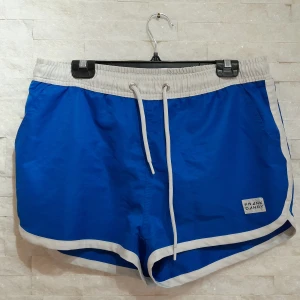 Blå badshorts från Frank Dandy - Snygga blå badshorts från Frank Dandy med vita detaljer och snörning i midjan. Klassisk sportig look med vit kant längs benen och elastisk midja. Perfekta för stranden eller poolen.