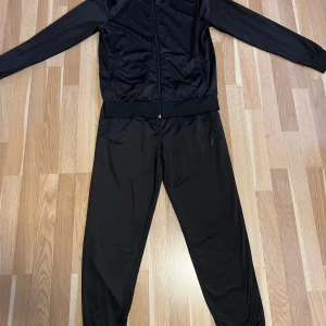 Svart träningsjacka från Emporio Armani EA7 - Säljer en svart träningsjacka från Emporio Armani EA7 med dragkedja framtill och hög krage. Jackan har en sportig look och EA7-logga på bröstet.  Snygga byxor till tröjan medföljer . Hela dressen är stel M .   Jätte fint skick !