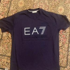 Mörkblå t-shirt från Emporio Armani EA7 - Säljer en mörkblå t-shirt från Emporio Armani EA7. T-shirten har en klassisk passform med korta ärmar och stort EA7-tryck på bröstet. Perfekt för dig som gillar stilrena märkeskläder. Pris kan diskuteras.