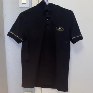 Svart t-shirt från EA7 Emporio Armani - Svart piké  från EA7 Emporio Armani med korta ärmar och knappar framtill. Guldiga detaljer på ärmsluten och EA7-logga på bröstet. Klassisk passform och tillverkad i bomull. Den här pikén är i storlek s men den passar även i M den är perfekt nu tiill en go och varm sommardag. Jag köpte dennna piken  för några månader sen och har inte kommit till användning därför gjorde jag  ett val att sälja den, Behövs de fler bilder är det bara att höra av sig. 