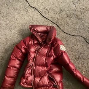 Vinröd dunjacka från Moncler - Säljer en vinröd dunjacka från Moncler med hög krage och dragkedja. Jackan har Moncler-logga på ärmen och quiltad design. Perfekt för kyliga dagar och riktigt snygg färg som sticker ut. priset kan diskuteras 