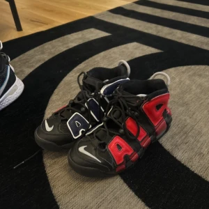 Nike Air More Uptempo sneakers i svart, rött och blått - Säljer ett par Nike Air More Uptempo sneakers med stora AIR-bokstäver i rött och blått på sidorna. Skorna har svart bas, vita detaljer och klassisk snörning. Perfekta för dig som vill sticka ut med en ikonisk streetwear-look.