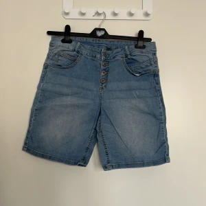 Blå jeansshorts med knappar från KappAhl - Snygga blå jeansshorts från KappAhl med fem knappar framtill och klassiska fickor både fram och bak. Modellen har en normal passform och är tillverkad i denim. Perfekta för sommardagar!