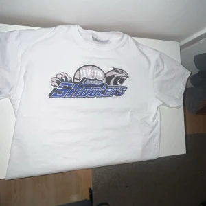 Vit t-shirt från Trapstar London Shooters - Säljer en vit t-shirt från Trapstar med stort tryck på bröstet där det står 'London Shooters' i blått och svart. T-shirten har rund hals och korta ärmar. Perfekt för dig som gillar streetwear och sportiga vibes.