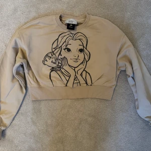 Beige croppad sweatshirt med Belle-tryck från Disney - Säljer en beige croppad sweatshirt från Disney med ett stort svart tryck av Belle och Chip från Skönheten och Odjuret på framsidan. Tröjan har långa ärmar och bred ribbad mudd i midjan. Använd en gång! superfin! 