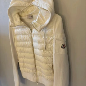 Vit pufferjacka med huva från Moncler - Säljer en snygg vit pufferjacka från Moncler med huva och dragkedja. Jackan har quiltade partier framtill, långa ärmar och en ficka med Moncler-logga på ärmen. Perfekt för kyliga dagar.