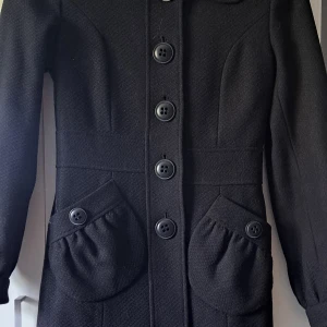 Svart kappa från Stockh LM med stora knappar - Säljer en svart kappa från Stockh LM med stora dekorativa knappar framtill och på fickorna. Jackan har markerad midja, långa ärmar och två fickor med lock. Klassisk och stilren design som passar till många tillfällen. Den är I jättefint skick. Ytterkläder består av 80% ull.