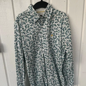 Vit blommig skjorta från Dressmann - Säljer en vit skjorta från Dressmann med grönt blommigt mönster. Skjortan har långa ärmar och klassisk krage. Insidan av manschetterna har diskreta svarta prickar. Perfekt för dig som vill sticka ut lite extra. 