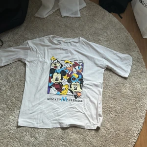 Vit t-shirt med Disney-tryck Mickey & Friends - Vit t-shirt med stort färgglatt tryck av Disney-karaktärerna Mickey Mouse, Pluto, Kalle Anka och Långben på framsidan. Klassisk passform med rund hals och korta ärmar. Perfekt för dig som gillar Disney och vill ha en lekfull stil.