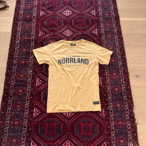 Gul t-shirt från SQRTN med Norrland-tryck - Gul t-shirt från SQRTN med svart text 'NORRLAND' på bröstet. Klassisk passform och rund halsringning. Perfekt för dig som gillar streetwear med norrländsk känsla.