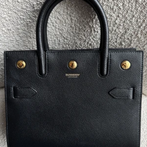 Svart handväska från Burberry - Säljer en stilren svart handväska från Burberry i kalvskinn med guldfärgade metalldetaljer. Väskan har två handtag och en klassisk, fyrkantig form. Avtagbar axelrem medföljer. Perfekt accessoar för dig som gillar tidlös design och lyxiga detaljer. Garanterar dess äkthet! kvitto finns ej. Mått: bredd 25,5 cm x längd 21,5 cm x bredd 9 cm