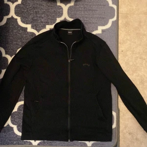 Svart zip-jacka från BOSS - Snygg svart jacka från BOSS med dragkedja framtill och diskret logga på bröstet. Jackan har hög krage och två fickor på sidorna. Perfekt för en stilren och enkel look.