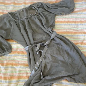 Ljusgrå klänning med knytband - Säljer en ljusgrå omlottklänning i tunn, luftig polyester med knytband i midjan. Klänningen har trekvartsärmar med resår och en lätt v-ringning. Perfekt för varma dagar eller som ett lager över badkläder.