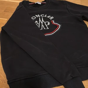 Svart sweatshirt från Moncler - Svart sweatshirt från Moncler med broderad logga på bröstet i vitt och rött. Tröjan har rund halsringning och långa ärmar med ribbade muddar vid ärmslut och nederkant. Perfekt för en stilren och sportig look.