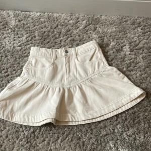 Vit volangkjol med jeanslook - Säljer en söt vit kjol med volang och jeansinspirerad design. Kjolen har knapp och dragkedja framtill samt markerad söm i midjan. Perfekt för dig som gillar en enkel men trendig stil. Aldrig använd 
