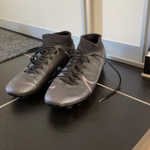 Svarta Nike Mercurial fotbollsskor - Säljer ett par svarta Nike Mercurial fotbollsskor med snörning och högt skaft. Skorna har en mönstrad ovandel i syntetmaterial och fasta dobbar för spel på gräs. Snygg design med diskreta silverdetaljer och Nike-logga på sidan. Finns en liten skada på vänster dojan vid fotleden men inget som påverkar prestandan enligt mig. På höger skon har en liten del av en dobb ramlat av som man kan se på sista bilden, återigen inget som påverkar prestandan så mycket de det inte är en viktig dobb