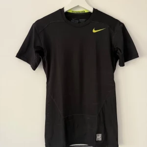 Svart Nike Pro Combat Hypercool t-shirt - Säljer en svart Nike Pro Combat Hypercool t-shirt med korta ärmar och gul logga framtill. Baksidan har ventilerande meshpaneler i gult och grått för extra andningsförmåga. Perfekt för träning och sport.