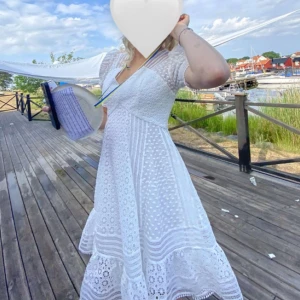 Abby dress By Malina - Superfin vit kortklänning med v-ringning, puffärmar och spetsdetaljer. Klänningen har markerad midja, volang nertill och vackert mönstrad spets i hela tyget. Perfekt för studenten och sommarens alla tillfällen!