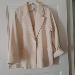 Beige kavaj med stora slag - Stilren beige kavaj med stora slag och två stora fickor framtill. Jackan har lång ärm och stängs med knappar. Perfekt för en clean och klassisk look.