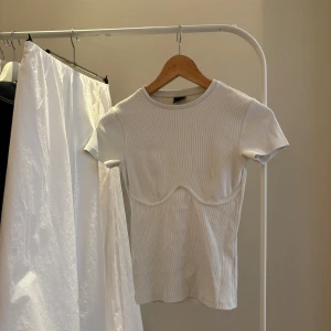 Vit ribbad topp från Gina Tricot - Säljer en vit ribbad topp från Gina Tricot med korta ärmar och rund halsringning. Toppen har en  sömdetalj som gör att den blir lite som en korsett. Perfekt att styla till jeans eller kjol!