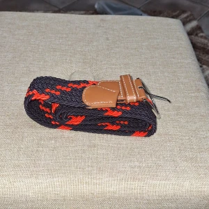 Svart och rött flätat bälte med bruna skinn-detaljer - Säljer ett snyggt flätat bälte i svart och rött med bruna skinn-detaljer vid spännet. Bältet har ett klassiskt metallspänne och passar perfekt till jeans eller chinos. Ger en cool touch till outfiten.