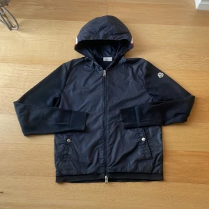 Svart jacka med huva från Moncler - Snygg svart jacka från Moncler med huva och dragkedja. Jackan har långa ärmar och ribbade muddar. Den har också två fickor med knappar framtill och en diskret logga på ärmen. Perfekt för en stilren look. INTE SAMMA INLÄGG 