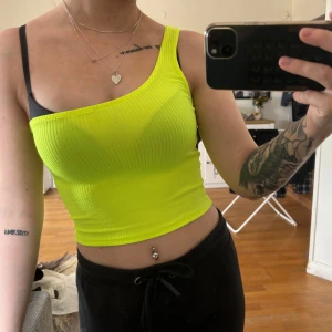 Neongul ribbad one shoulder topp - Säljer en neongul ribbad topp med one shoulder-design. Toppens material är stretchigt och sitter tight. Perfekt för dig som vill sticka ut med en färgstark och trendig look.