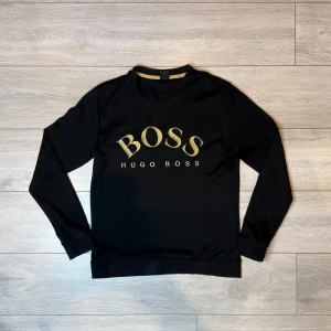 Svart sweatshirt från Hugo Boss med guldtryck - Gold in black Hugo boss crewneck. Condition 7/10 (few flaws are seen on the pictures, but overall not noticeable) Size M For more information or details feel free to contact us privately.
